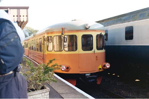 An orange and yellow railcar sits in a station which is mostly obscured by someone standing just out of shot.  A dull grey train sits on the other side.