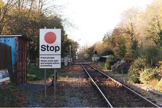 Looking down the tracks towards a road crossing.  A stop sign in the foreground lists instructions to train drivers.  The sun, streaming between trees, makes the sky glow white.