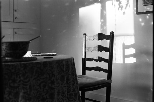 an empty chair at a table with a bowl.  the glowing shape of light from a window can be seen on the wall and chair.  specks of light dance across the walls.