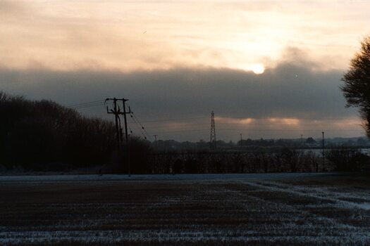A view over stubbly, snowy fields, dark woods, and utility poles of a pale sky.  The sun burns partially through a dark cloud.