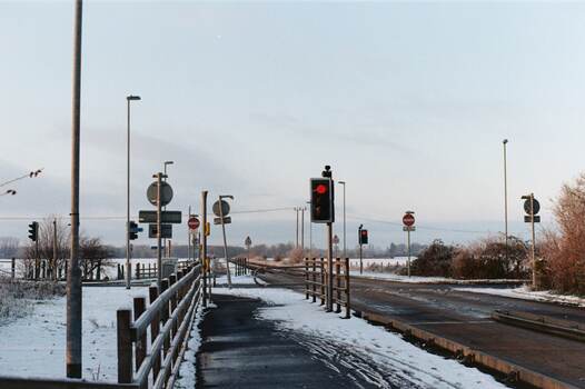 An empty junction adorned with road furniture and surrounded by flat, white fields.  Black tarmac is visible under the snow.  The traffic lights are red.