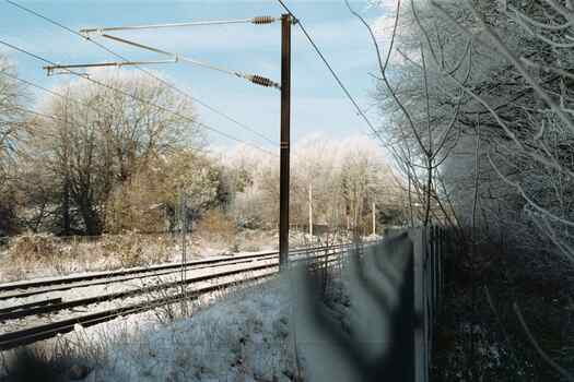 A view over a fence onto a railway, taken from the shade under some frosty trees.  The snowy railway is brightly lit by the sun, under a clear blue sky.