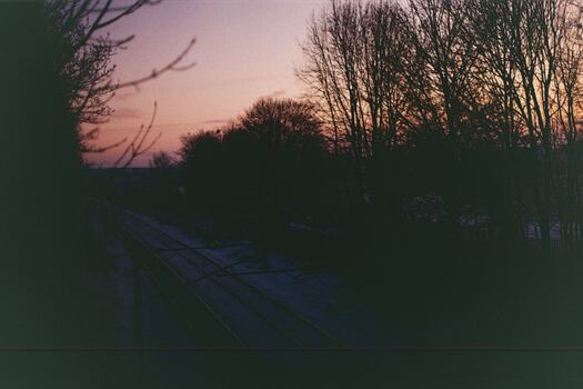 A dark, moody photo of a railway, taken from a bridge over it, at dusk.  The ground is nearly lost in silhouette.  The sky is a soft pink and purple gradient.